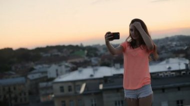 Turuncu t-shirt ve şort güzel kadın gün batımında gülümseyerek telefon kullanarak selfie alarak ve çatıdan cityscape görünümü zevk