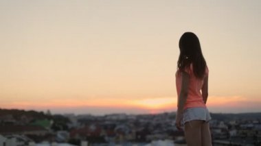 Güzel gülüşlü esmer kadın fotoğrafçı pozu veriyor. Gün batımında şehir manzaralı yüksek bir platformda duruyor.