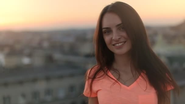 Sexy meisje met lang haar draaien rond en glimlachend op camera buiten, wazig stad bij zonsondergang op achtergrond — Stockvideo