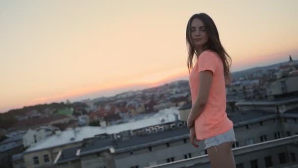 Hermosa mujer sonriente bailando con el pelo largo que sopla y levantando brazos en la puesta del sol en el techo de apartamentos — Vídeos de Stock