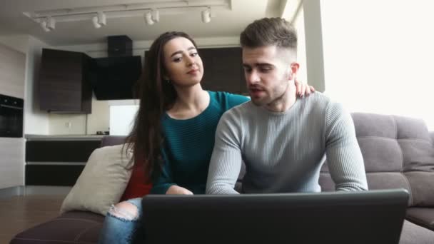 Belo casal sentado em um sofá, jovem verificando seus e-mails no laptop e falando com a namorada, beijando-a nos lábios . — Vídeo de Stock