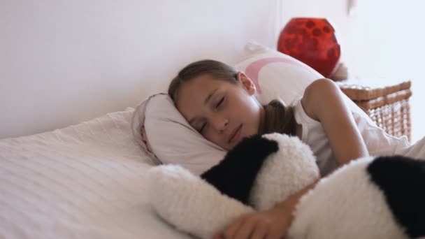 Очаровательная маленькая девочка отдыхает в постели с мягкой игрушкой и смотрит в камеру крупным планом — стоковое видео