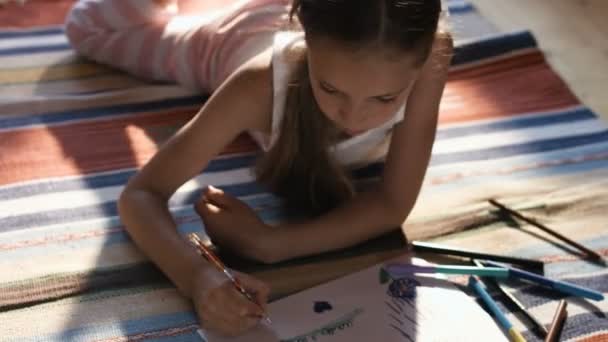 Chica encantadora acostada en el dibujo de la alfombra con lápices de colores en el papel — Vídeos de Stock