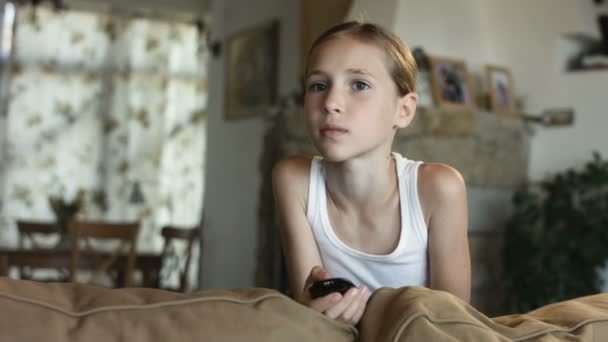 Retrato de la adorable niña sentada en el sofá y sosteniendo en su mano un control remoto mientras ve dibujos animados en la televisión — Vídeos de Stock