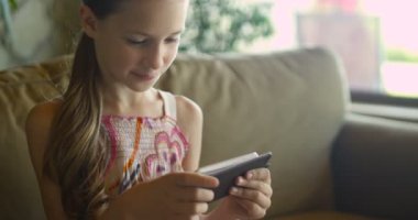 Oyun oynarken veya oturma odasında beyaz Koç oturan tablet app kullanarak küçük kız. Rahat beyaz ev kanepede oturan ve akıllı telefon ekranına bakarak güzel çocuk kız