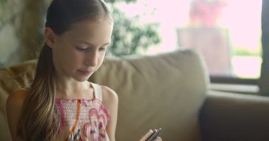 Oyun oynarken veya oturma odasında beyaz Koç oturan tablet app kullanarak küçük kız. Rahat beyaz ev kanepede oturan ve akıllı telefon ekranına bakarak güzel çocuk kız