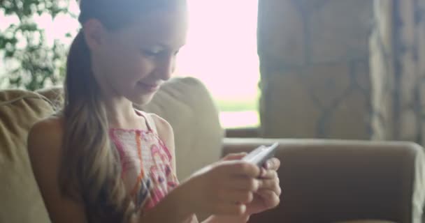 Kleines Mädchen, das Spiele spielt oder eine App auf dem Tablet nutzt, sitzt auf einer weißen Kutsche im Wohnzimmer. schöne junge Mädchen sitzt auf gemütlichen weißen Sofa zu Hause und schaut auf Smartphone-Bildschirm — Stockvideo