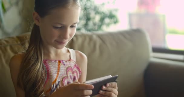 Niña jugando juegos o utilizando la aplicación en la tableta sentado en el entrenador blanco en la sala de estar. Hermosa niña sentada en el acogedor sofá blanco en casa y mirando la pantalla del teléfono inteligente — Vídeo de stock