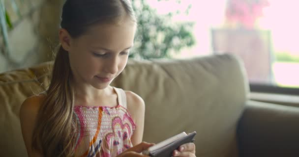 Menina brincando ou usando o aplicativo no tablet sentado no treinador branco na sala de estar. Menina bonita criança sentada no sofá acolhedor casa branca e olhando para a tela do smartphone — Vídeo de Stock