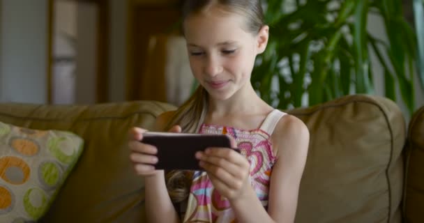 Маленька дівчинка грає в ігри або використовує додаток на планшеті, сидячи на білому тренері у вітальні. Красива дівчинка сидить на затишному білому домашньому дивані і дивиться на екран смартфона — стокове відео