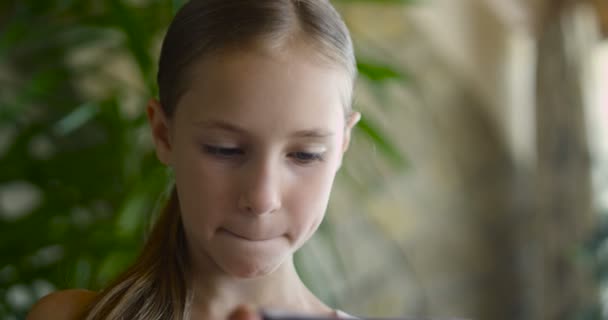 Retrato de cerca de una niña pequeña sosteniendo y usando un teléfono celular inteligente enfocado mientras está de pie en una sala de estar en casa, interior . — Vídeo de stock