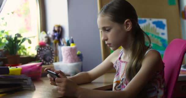 Маленькая улыбающаяся школьница, сидящая со смартфоном и играющая в игры за столом в детской — стоковое видео
