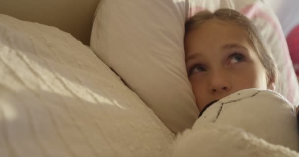 Linda niña bostezando en pijama acostada en una cama debajo de una manta blanca en la mañana soleada — Vídeo de stock