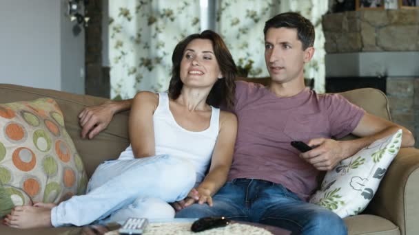 Feliz y encantadora pareja joven en casa en el sofá viendo la televisión juntos, hombre con control remoto — Vídeos de Stock