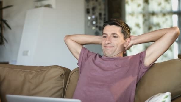 Jeune homme attrayant utilise un ordinateur portable et souriant tout en étant couché avec les mains derrière la tête sur le canapé à la maison — Video