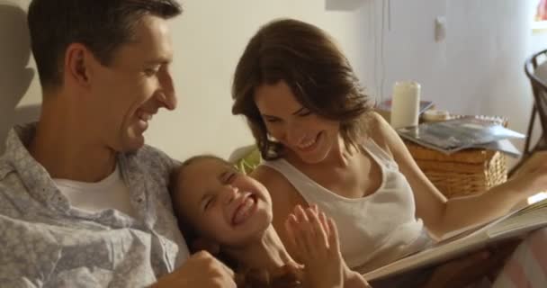 Madre, padre e hija con pijama leyendo un libro juntos acostados en la cama — Vídeo de stock