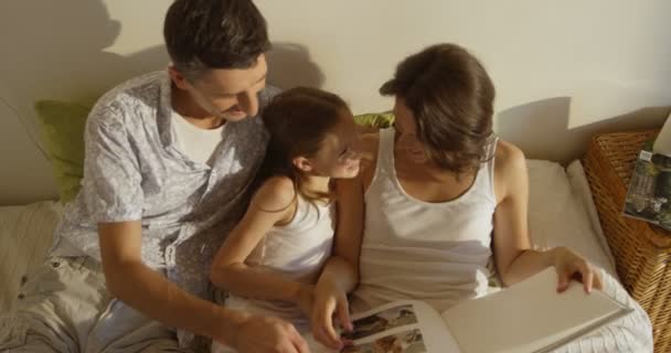 Mor, far och dotter klädd i pyjamas läser en bok tillsammans om i sängen — Stockvideo