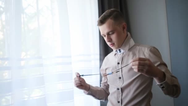 Elegante novio de moda joven en camisa beige vestidor pajarita de pie cerca de la ventana — Vídeos de Stock