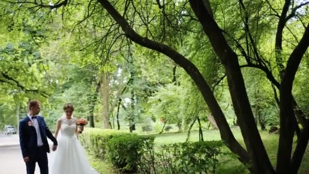 Просто подружня пара йде стежкою в парку. Наречена тримає весільний букет з троянд — стокове відео