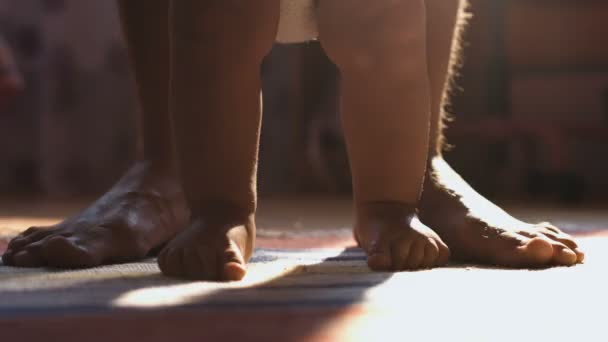 Gros plan de bébé garçon marchant ses premiers pas avec papa derrière, dans une pièce ensoleillée — Video