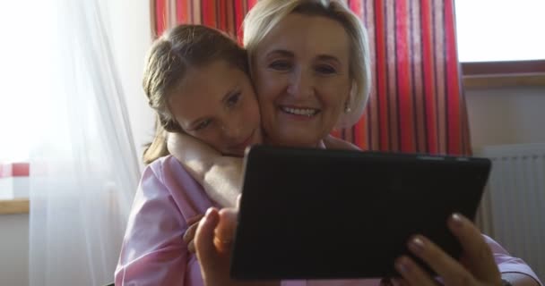 Семья, поколение, технологии и люди - улыбающиеся внучка и бабушка с планшетным компьютером, сидящие дома на диване — стоковое видео