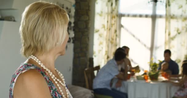 Charmig äldre kvinna poserar på kameran och familjen sitter vid dinning bord i bakgrunden — Stockvideo