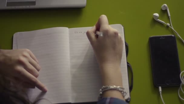 Mulher mão escrevendo em notebook em branco aberto com telefone celular e fones de ouvido em uma mesa, vista superior — Vídeo de Stock