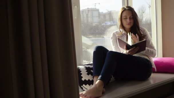 Encantadora joven con estilo leyendo un libro en el alféizar de la ventana y mirando a través de la ventana — Vídeo de stock