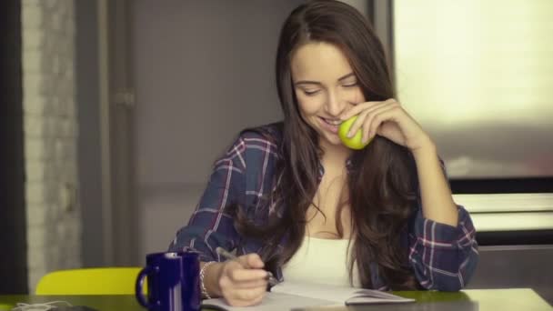 Sorridente bella giovane donna seduta a scrivere nella sua sala da pranzo a tavola tenendo mela verde come spuntino sano — Video Stock