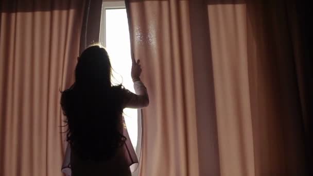 Atrakcyjna młoda kobieta otwiera zasłony w zwolnionym tempie to patrząc na wschód stoi blisko okna w swoim domu i cieszy się z widokiem na miasto z wysokości — Wideo stockowe