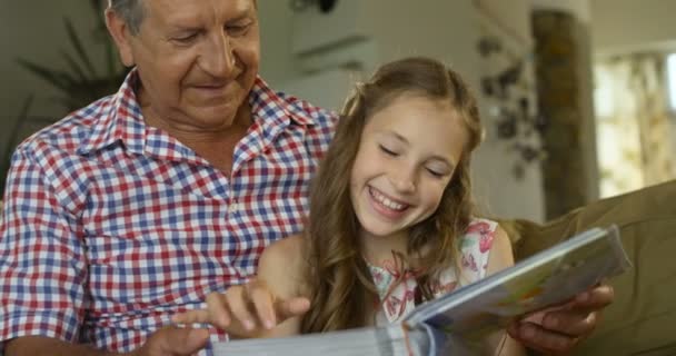 Söt flicka sitter i knät på sin farfar och ser glatt tillsammans på ett fotoalbum — Stockvideo