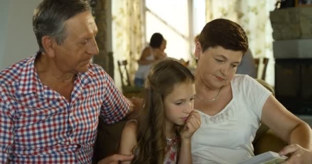 Abuelo sonriente, nieta y abuela con libro o álbum de fotos sentados en el sofá en casa — Vídeos de Stock