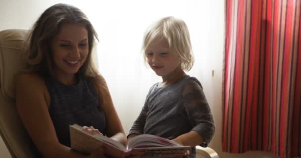 Красива блондинка мама читає книгу дочці, сидячи в кріслі — стокове відео