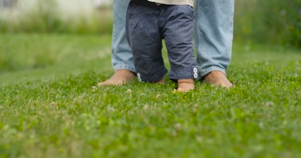 Padre aprende a caminar al aire libre. Bebé primeros pasos en la hierba, cámara lenta . — Vídeos de Stock