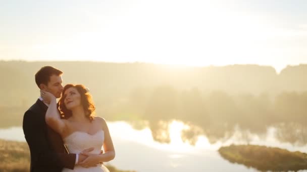 Jong paar in liefde bruid en bruidegom op de achtergrond van zonsondergang, rechtzet haar backlight, veld van tarwe, zacht en romantisch, witte jurk, das, natuur, schot in slow motion close-up — Stockvideo