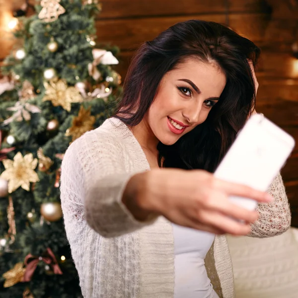 Personas, vacaciones y el concepto de la tecnología - hermosa mujer morena en suéter blanco tomando foto selfie por teléfono inteligente sobre el árbol de Navidad y el fondo de la sala de estar — Foto de Stock