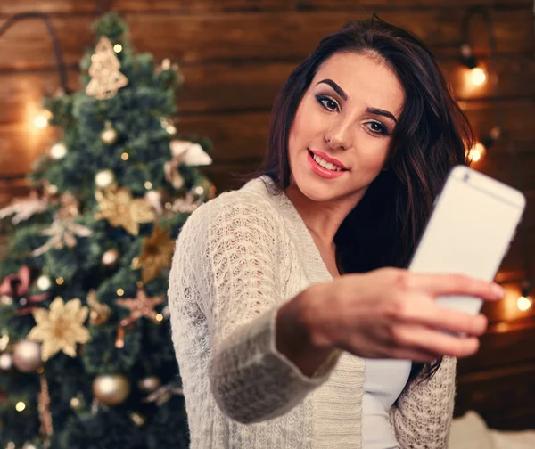 people, holidays and technology concept - beautiful brunette woman in white  sweater taking selfie picture by smartphone over christmas tree and living room background