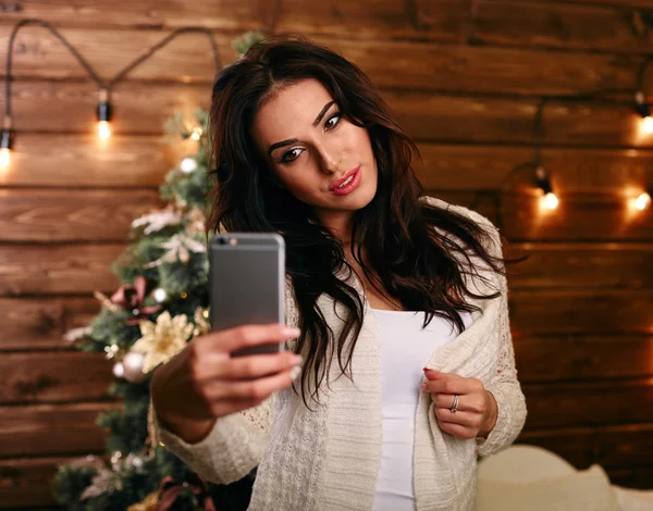 people, holidays and technology concept - beautiful brunette woman in white  sweater taking selfie picture by smartphone over christmas tree and living room background