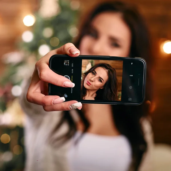 people, holidays and technology concept - beautiful brunette woman in white  sweater taking selfie picture by smartphone over christmas tree and living room background