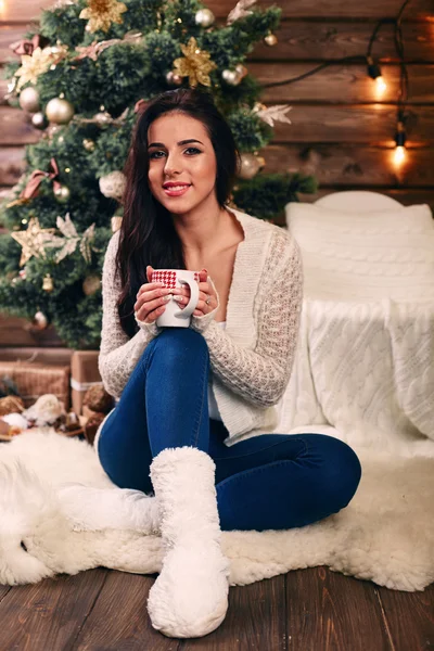 Schöne junge Frau trinkt Tee am Weihnachtsbaum. schöne Mädchen feiert Weihnachten mit einer Tasse Kakao vor Baum über Wohnzimmer — Stockfoto