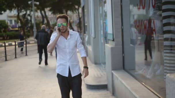 . Güneş gözlüğü takan, şehir merkezinde yürüyen ve telefonla konuşan genç adam. 20'li yıllarda kentsel erkek. — Stok video