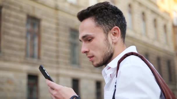 Tipo atractivo mirando en la pantalla de un teléfono y sonriendo. Hombre positivo en sus 20 años utilizando el teléfono inteligente al aire libre — Vídeos de Stock