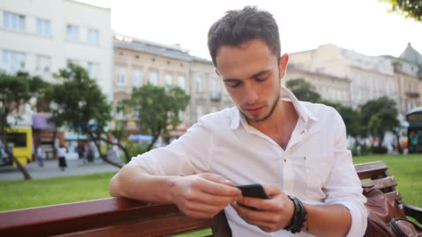 Hipster usando su dispositivo para crear mensajes. Tipo guapo sentado en una plaza charlando en el teléfono inteligente . — Vídeo de stock