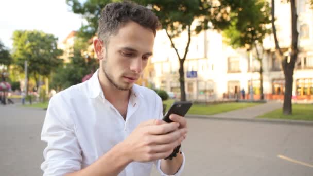 Brownhaired młody człowiek za pomocą smartphone na zewnątrz. Facet, wpisując na telefon w parku miejskim. Zbliżenie: mężczyzna ręce stukanie na urządzeniu, przewijanie, analytics, diagramy, wykresy. — Wideo stockowe