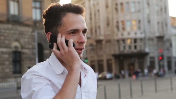 Ragazzo bello e positivo che parla attivamente al cellulare all'aperto durante il tramonto nel mezzo della città europea . — Video Stock