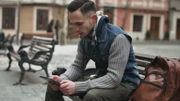 Knappe jonge man zittend op een bank in het centrum van de Europese stad met behulp van een Tablet. Kerel speelt online games op zijn mobiele apparaat. — Stockvideo