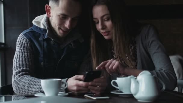 Joven hombre y mujer sentado en el café y mirando en la pantalla de la pareja feliz smartphone jugando juegos en el dispositivo y sonriendo el uno al otro — Vídeos de Stock