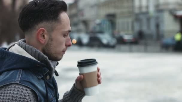 Young male is drinking takeaway coffee outdoors and waiting for someone. Attractive man sipping coffee in the city center while looking forward to meet his friend. — Stock Video