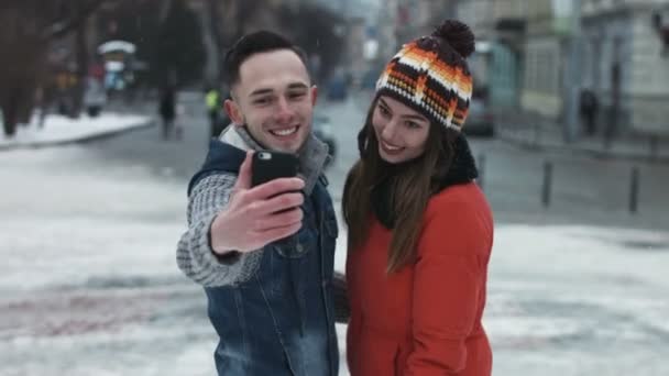 Giovane coppia attraente fare facce sulla macchina fotografica. Uomo e donna scattare fotografie utilizzando smartphone. Ragazzo e ragazza fanno selfie all'aperto nel bel mezzo del centro storico europeo mentre nevica . — Video Stock