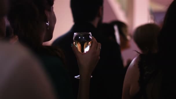 Ung attraktiv kvinna stående tillbaka till kameran och hålla ett glas vitt vin på festen och titta på i ett skede där en föreställning pågår. — Stockvideo